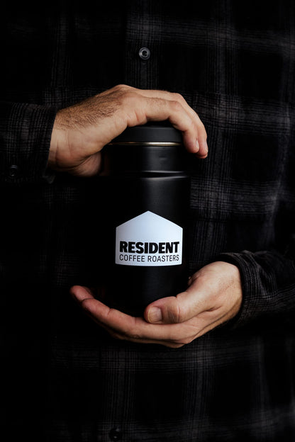 A person in a black plaid shirt holds a matte black insulated container with a white house-shaped logo that reads 'RESIDENT COFFEE ROASTERS'.