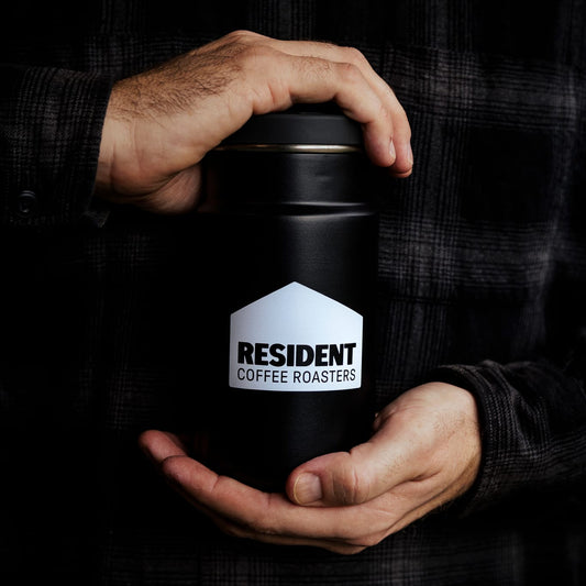 A person in a black plaid shirt holds a matte black insulated container with a white house-shaped logo that reads 'RESIDENT COFFEE ROASTERS'.